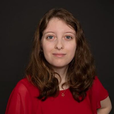 Elyse Hoffman Author Profile Image with wavy shoulder-length brown hair and wearing a red top, looking straight at camera against a dark background