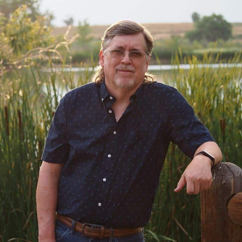 R. Weir author leaning on a fence post in front of a weeded pond with hair pulled back and a dark shirt