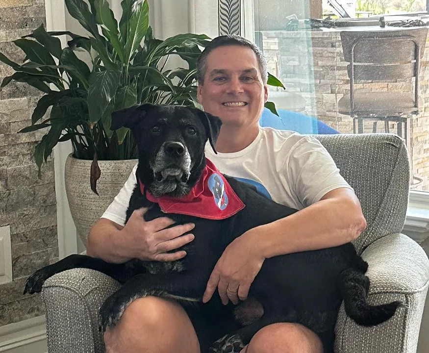 “Karl Riemensperger, smiling while seated in a light gray armchair, hugs his black rescue dog Gunner. Gunner wears a red bandana with a blue patch. They’re indoors, with a large leafy plant and sunlit windows in the background.”