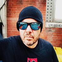 Golden Blood: A Vampire Story by Tim Vee ~ $25 Gift Card Available #Horror #Thriller #Vampire #DarkFantasy @GoddessFish @_Tim_Vee_ 7 Author Tim Vee wearing dark sunglasses, a black knit beanie, and a black T-shirt, standing against a red-brick industrial backdrop.