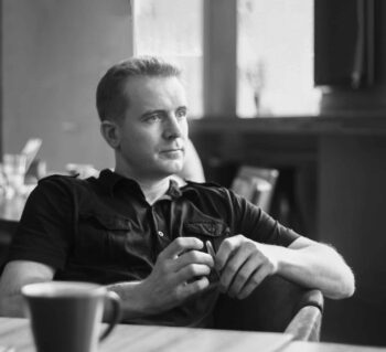 Black & white image of a man in a dark polo shirt sitting at a table with a cup of coffee and a pensive look on his face