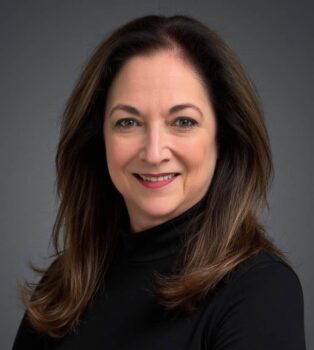 Professional headshot of author Margaret Izard. She has long brown hair with soft highlights, is wearing a black turtleneck, and is smiling against a gray backdrop.