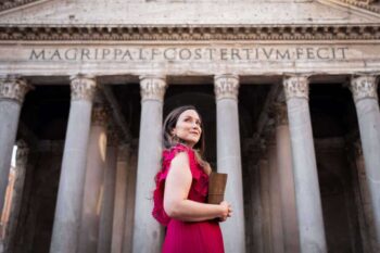 Author Flavia Brunetti wearing a red dress and holding a book, standing in front of a historic building. Promotional photo for her YA fantasy novel, The Web of Time.