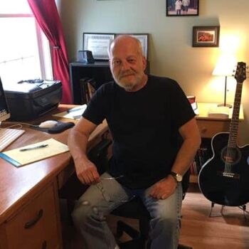 Gregory F. DeLaurier seated at his desk in casual clothes, writing on a yellow notepad with a pen, a guitar resting nearby—capturing the creative spirit behind his novel Renewals.