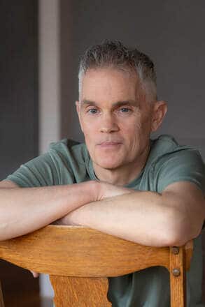 Author Daniel E. Chambrello leaning on the back of a chair with his arms crossed.