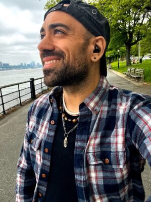 Author Benjamin Ryan profile picture, flannel shirt, backwards, black baseball cap, with a riverwalk background