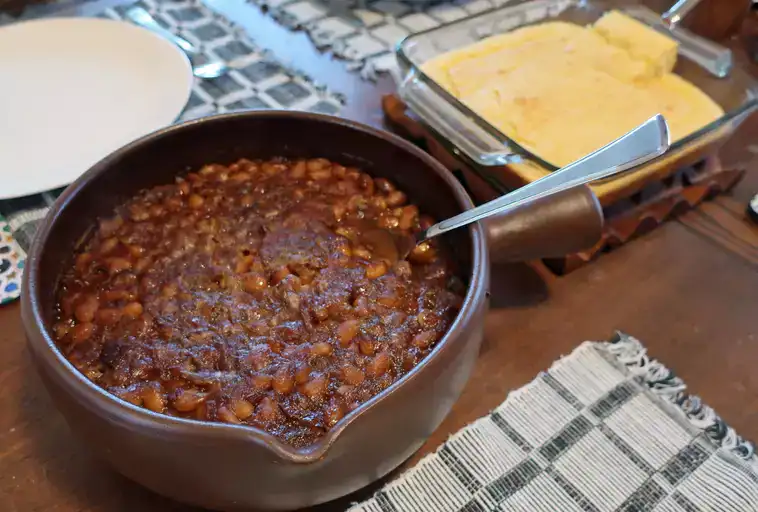 Chef John's Classic Boston Baked Beans in a crock sitting beside a dish of cornbread