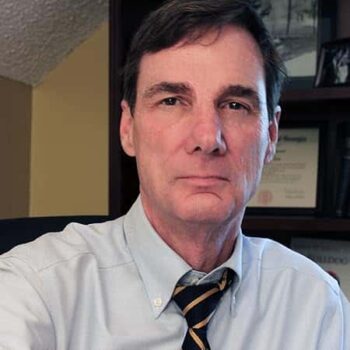 Headshot of author David Burnett. He wears a light blue shirt and striped tie, seated in an office with books and degrees visible behind him. Used for author bio and promotional materials for “Child of a Swan.”