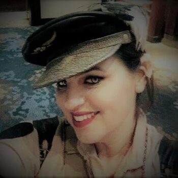 Author Kacey Ezell smiles in a vintage-style military cap and uniform, with bold eyeliner and red lipstick. She’s in a warmly lit indoor setting with a patterned blue carpet in the background.