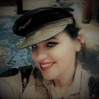 Magelight by Kacey Ezell – Romantic Fantasy Book Review, Excerpt & $25 Giveaway 14 Author Kacey Ezell smiles in a vintage-style military cap and uniform, with bold eyeliner and red lipstick. She’s in a warmly lit indoor setting with a patterned blue carpet in the background.
