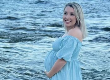 Author Larissa Pemberton stands by the ocean in a light blue off-shoulder dress, smiling warmly with one hand on her baby bump. The waves behind her reflect the gentle light, creating a peaceful and joyful atmosphere.