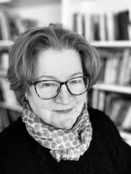 Black-and-white headshot of author Litty Williams, wearing glasses and a polka-dotted scarf, smiling slightly. Bookshelves are softly blurred in the background.