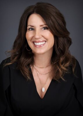 Author Michele Clarizio smiling in front of a neutral gray background. She has long dark brown hair, wears a black top with a gold necklace, and projects warmth and confidence.