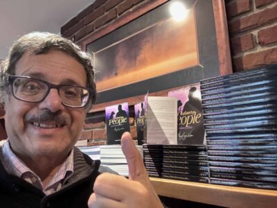 Author Naguib Sami Kerba smiling in front of a display of Ordinary People, Extraordinary Lives books. He gives a thumbs-up next to neatly stacked copies.”