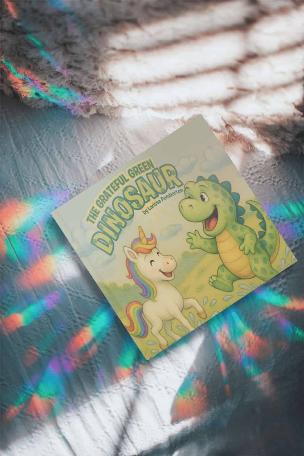 Photo of “The Grateful Green Dinosaur” picture book on a soft surface with rainbow light reflections. Shows cheerful illustrated cover featuring a unicorn and dinosaur, highlighting themes of gratitude and positivity.