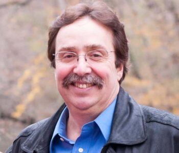 Author B.T. Polcari smiles in an outdoor autumn setting. He wears glasses, a blue collared shirt, and a black leather jacket. His brown hair and mustache give him a friendly, approachable appearance.