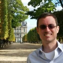 Author D. H. Schleicher smiling outdoors in a tree-lined park with a historic chateau-style building in the background. He is wearing sunglasses and a collared shirt.