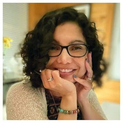 Spotlight: The Passenger by Joie Lesin – A 1940s Ghost Story of Love, Loss, and Second Chances 12 Portrait of Joie Lesin, award-winning Minnesota-based author of The Passenger. She is smiling, wearing glasses, with curly hair, resting her chin on her hands in a warmly lit indoor setting.
