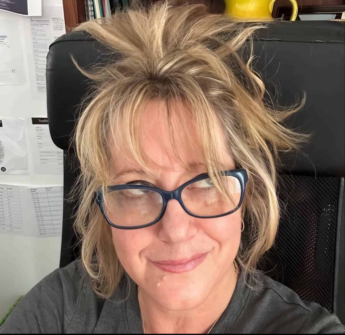 Author photo of LS Delorme. She is smiling with tousled blonde hair and blue glasses, seated in a black chair with papers pinned to the wall behind her. Warm, playful expression.