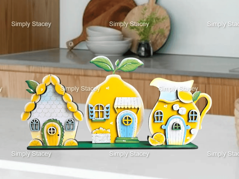 Lemon-themed wooden DIY craft kit from SimplyStacey on Etsy. Features three whimsical pieces: a cottage with lemon trim, a house shaped like a lemon, and a lemonade pitcher cottage with sugar cube details. Displayed on a kitchen counter with bright summer tones.