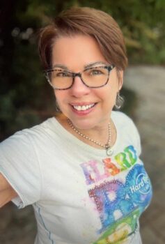 Author photo of Lori Matsourani, smiling outdoors while wearing glasses, a rainbow Hard Rock Café T-shirt, and a silver necklace.