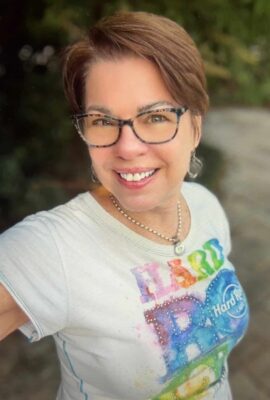 Author photo of Lori Matsourani, smiling outdoors while wearing glasses, a rainbow Hard Rock Café T-shirt, and a silver necklace.