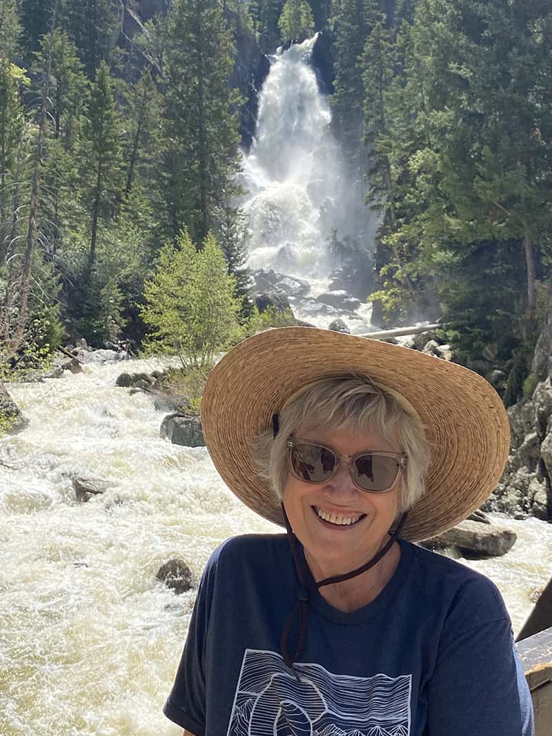 Author photo of Meg Benjamin smiling outdoors in front of a waterfall and pine forest. She wears sunglasses, a wide straw hat, and a navy T-shirt.
