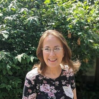 Rosie Russell, author and illustrator of children’s books, smiling outdoors in front of lush green foliage. She is wearing glasses and a black floral top with a white lace pocket detail.