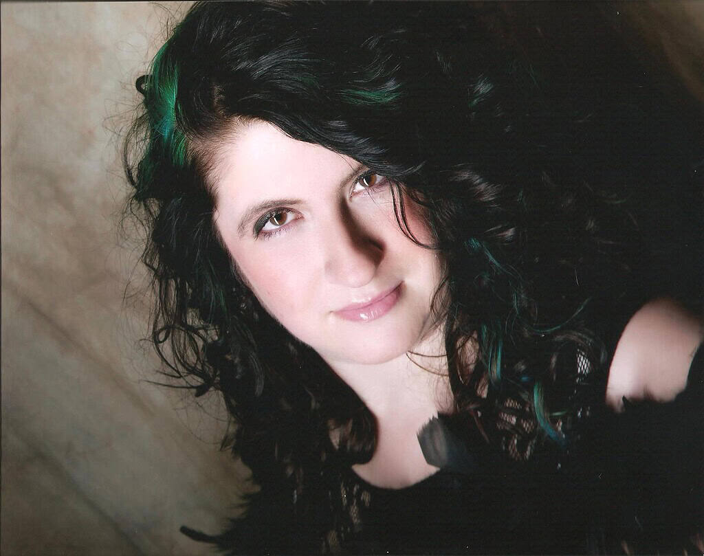 Author Tanith Davenport smiling in a close-up headshot; long dark curly hair with green highlights; black lace top; neutral backdrop.