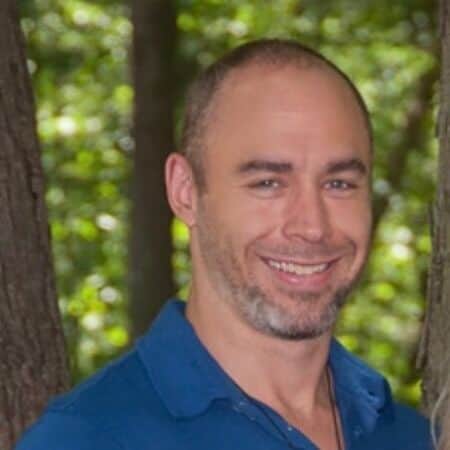 Author photo of Alex Zenk, smiling outdoors in a wooded background, wearing a blue collared shirt.