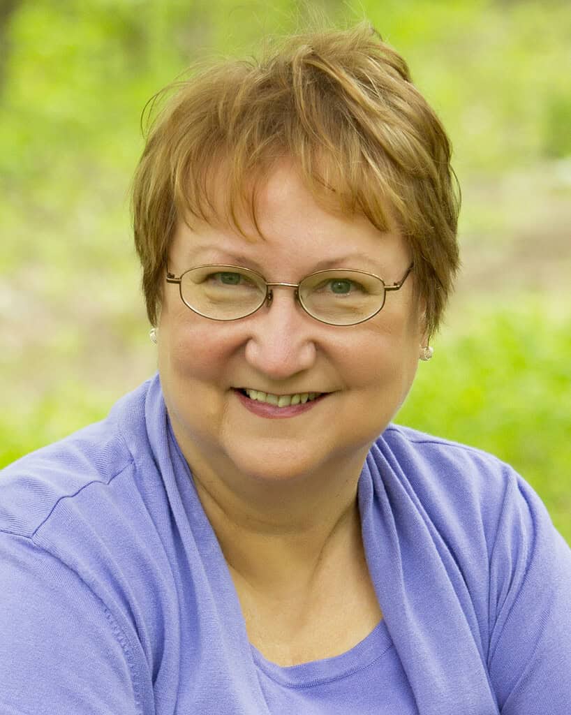 Author photo of Joyce Proell, co-author of Birds, Puppies, and Murder. Smiling woman in glasses and purple top, outdoors.