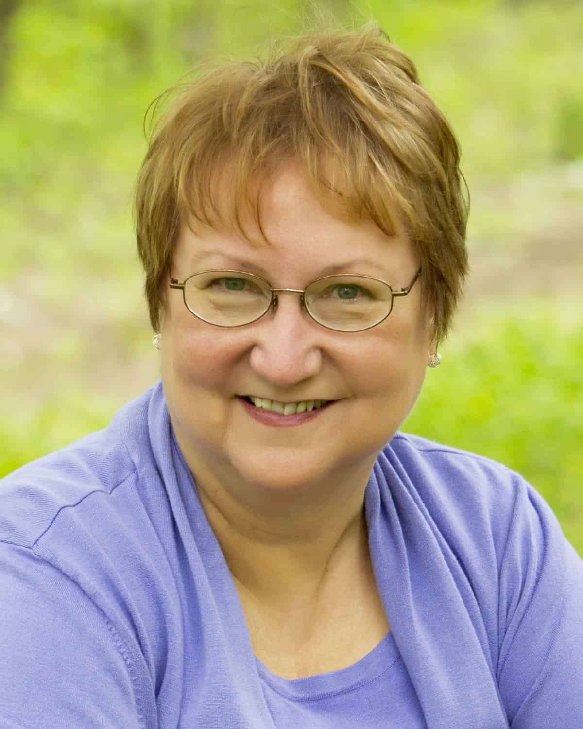 Birds, Puppies, and Murder by Joyce Proell & Brenda Whiteside | Cozy Mystery Review Tour 15 Author photo of Joyce Proell, co-author of Birds, Puppies, and Murder. Smiling woman in glasses and purple top, outdoors.