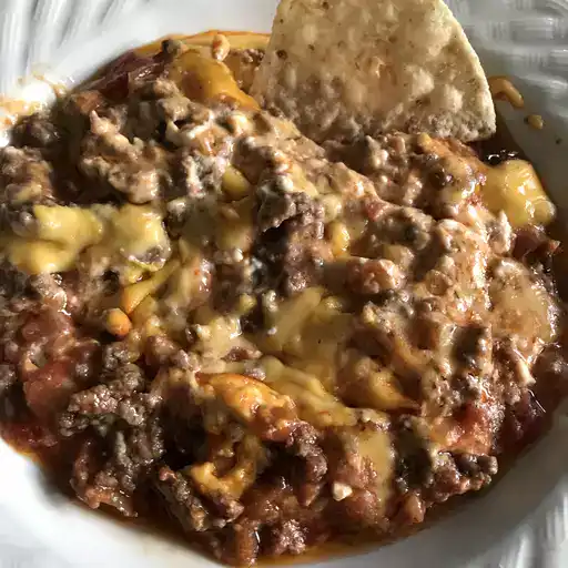 “Close-up of a hearty cheesy beef dip served in a white bowl, topped with melted cheddar and seasoned ground beef, with a tortilla chip ready for dipping—perfect for game day or parties.”