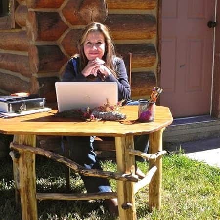 Author Cricket Rohman sitting outdoors at a rustic log cabin table with her laptop, smiling as she writes her next romantic mystery.