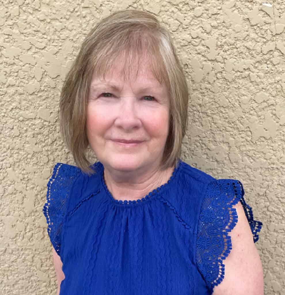 Author Janette C. Andrejowich, smiling against a neutral textured wall, wearing a royal-blue blouse with lace-trimmed sleeves