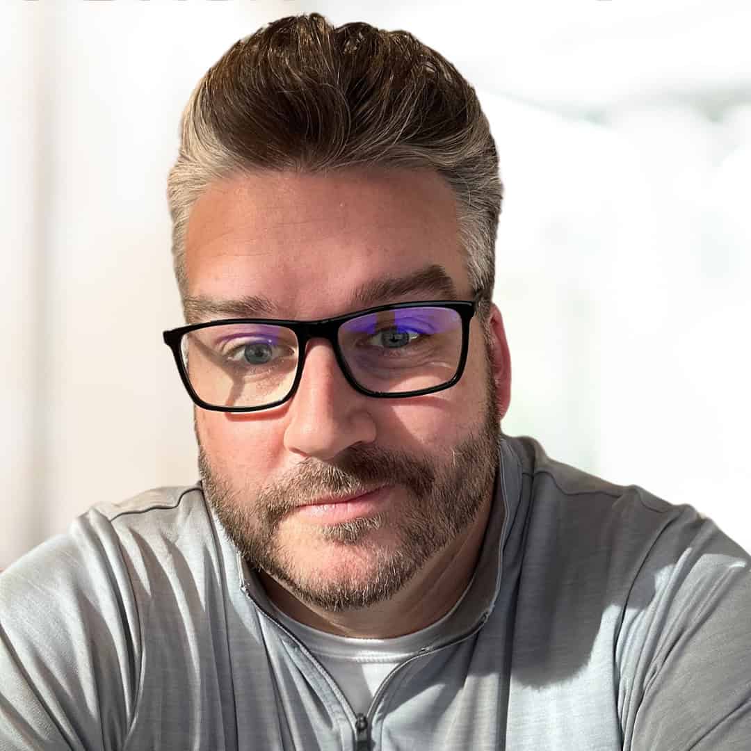 Author photo of Marc Macdonald. He has short styled hair, glasses, and a trimmed beard, wearing a light zip-up shirt, smiling slightly in a softly lit indoor setting