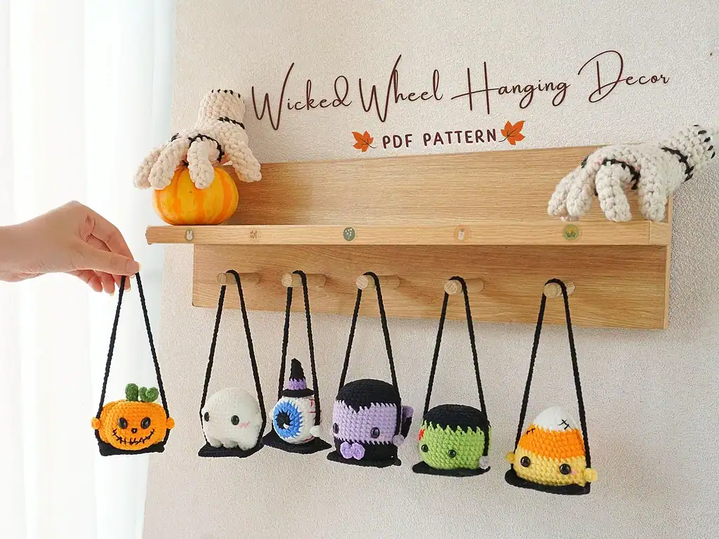 “Handmade Halloween-themed crochet hanging decor displayed on a wooden wall rack. Features a crocheted pumpkin, ghost, eyeball, witch, Frankenstein, and candy corn characters swinging from black yarn loops, with a small pumpkin and two crocheted skeletal hands perched on top. Text reads ‘Wicked & Wheel Hanging Decor – PDF Pattern.’”