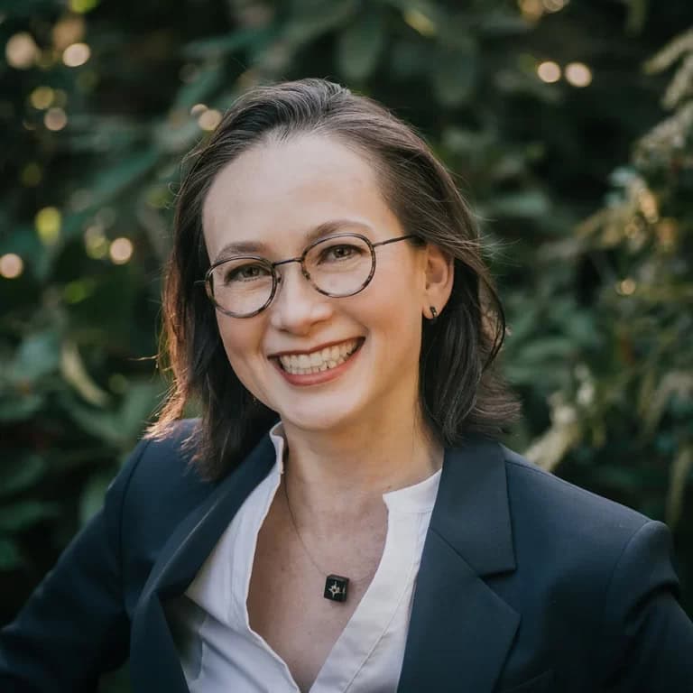 Ya-Ling-Liou-wearing a white blouse with a black jacket, dark hair, glasses and a open smile in front of tree background