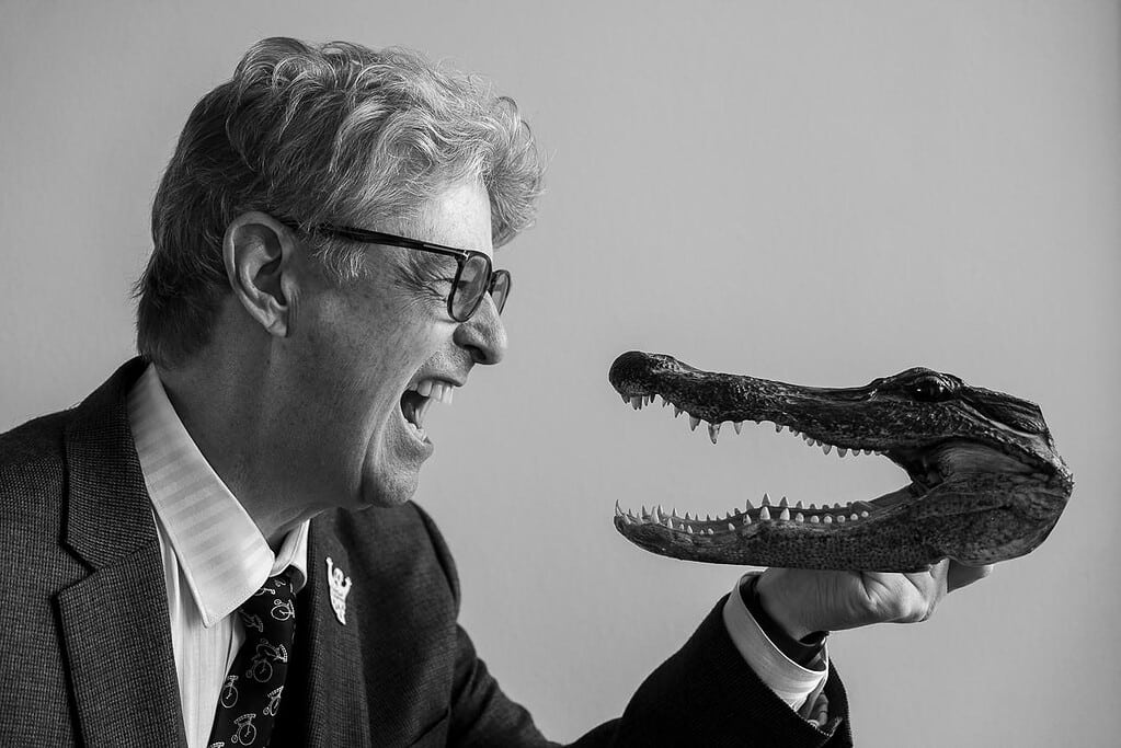 “Black-and-white portrait of author Mark Nutter playfully facing an open alligator skull prop, profiles meeting as he smiles; quirky, humorous author photo.”