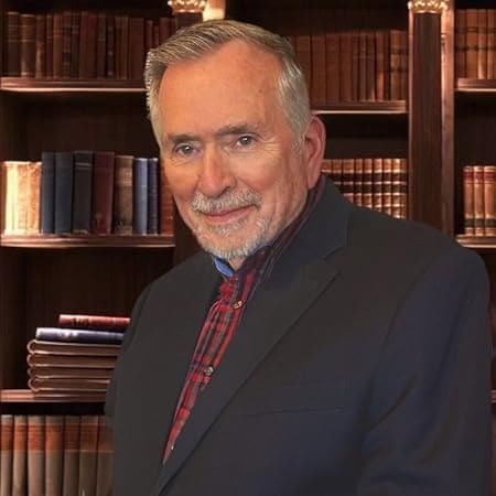 Author photo of Travis Short in a dark blazer with red plaid shirt, smiling in front of a bookcase filled with classic books.