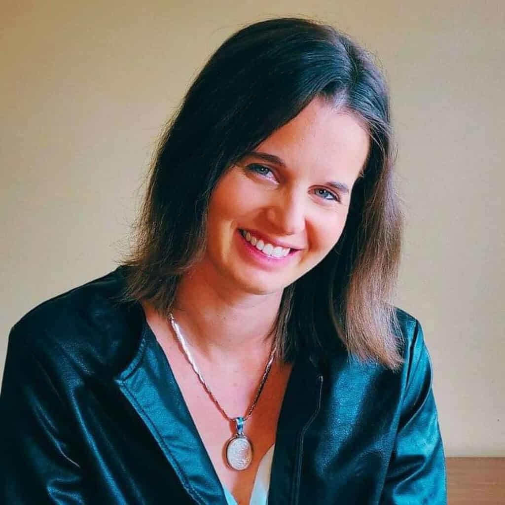 Author photo of Amber Leigh Williams smiling in a dark jacket with a silver pendant necklace against a neutral background. Used for Goddess Fish Promotions Review Tour materials for “Madame Rebelle.”
