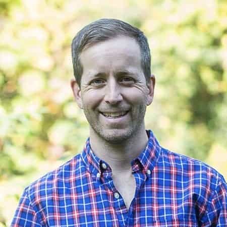 Author Andrew D. Thompson smiling outdoors in a blue and red plaid shirt with blurred greenery background. Thompson is the author of A High-Performing Mind.