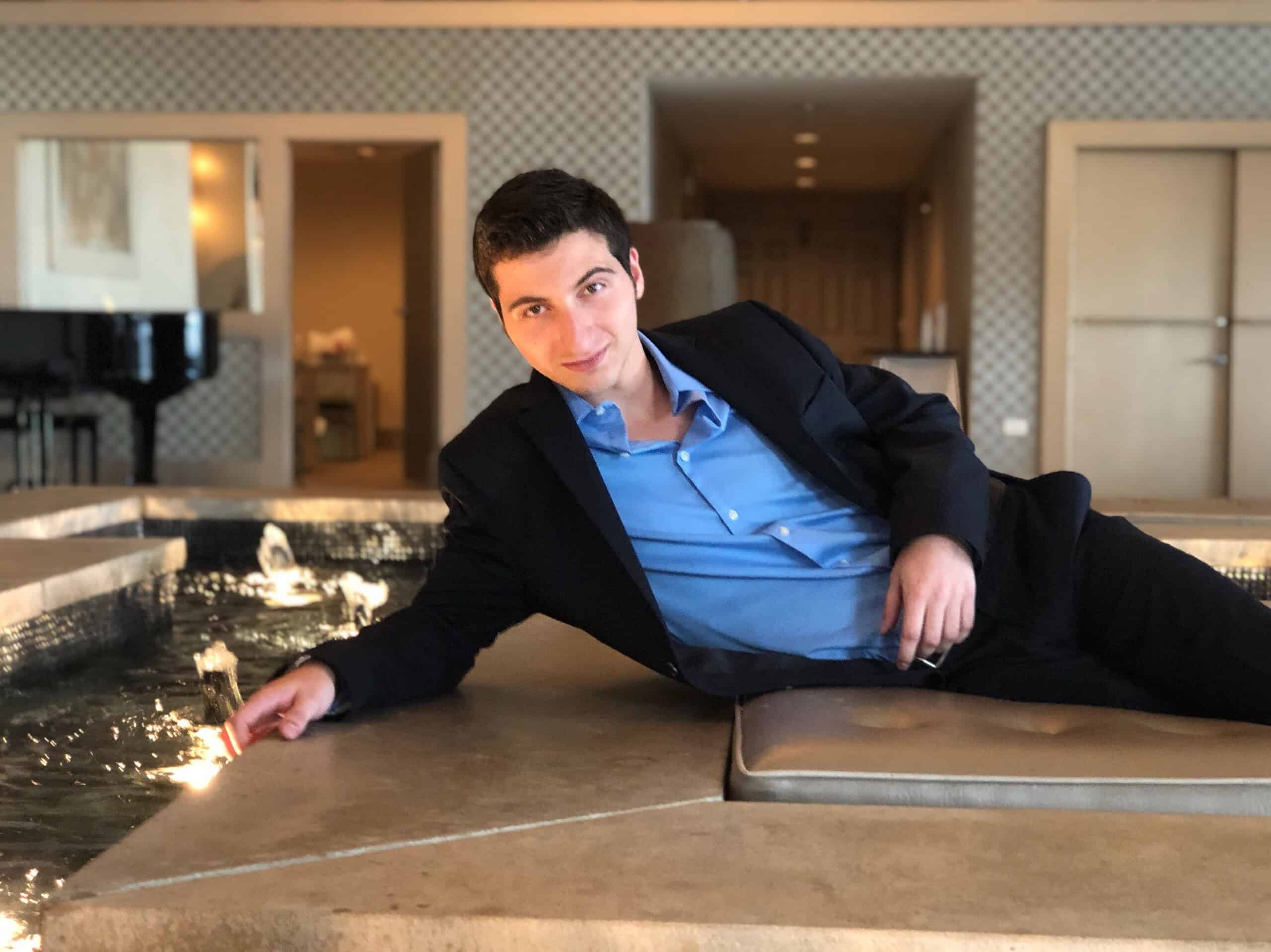 The Quest For Freedom by Matthew Devitt – Epic Fantasy Book Review 4 Author Matthew Devitt reclining beside a fountain in a dark suit with a light blue shirt, smiling at the camera in a warmly lit indoor setting.