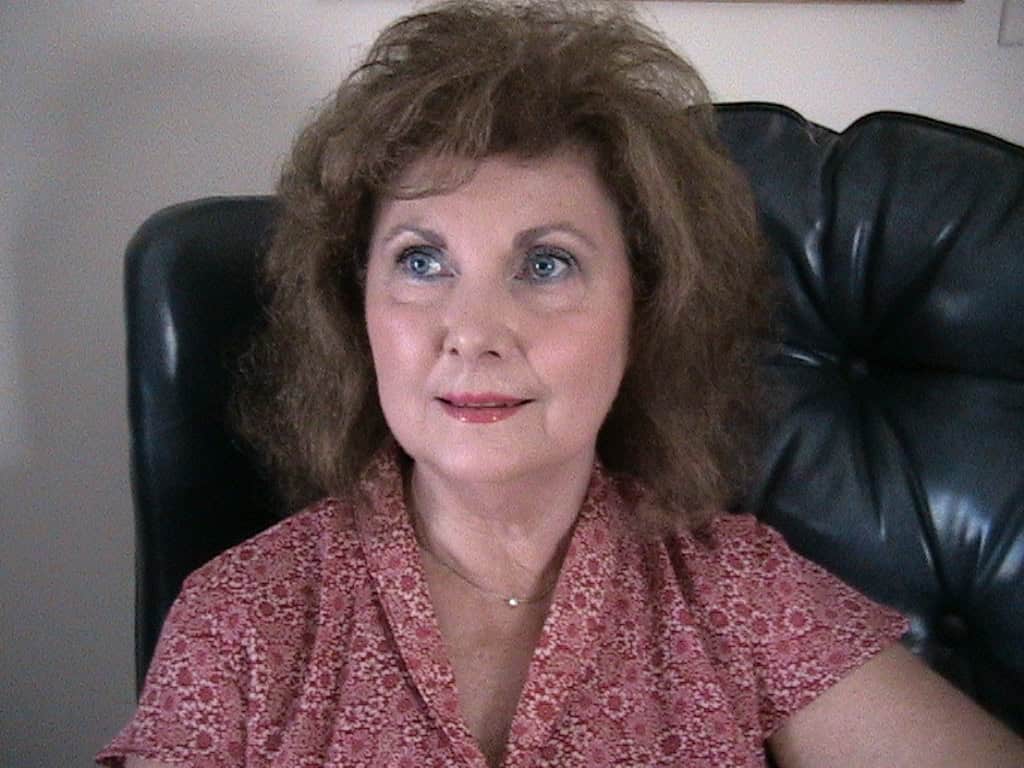 Author photo of Barbara Casey, writer of Shyla's Initiative. She is seated in a dark chair, wearing a patterned red blouse, with light brown hair and a calm, thoughtful expression