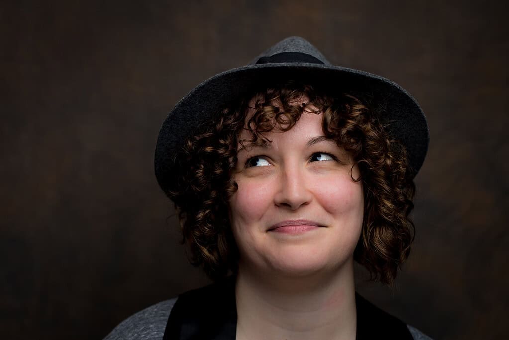 Author photo of D. Lieber. A smiling woman with curly brown hair wears a gray hat and looks playfully upward against a dark background.