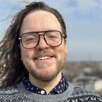 Author photo of Jeffrey Haskey-Valerius. He is smiling outdoors, wearing glasses and a patterned sweater over a collared shirt, with long hair flowing in the wind against a bright sky backdrop.