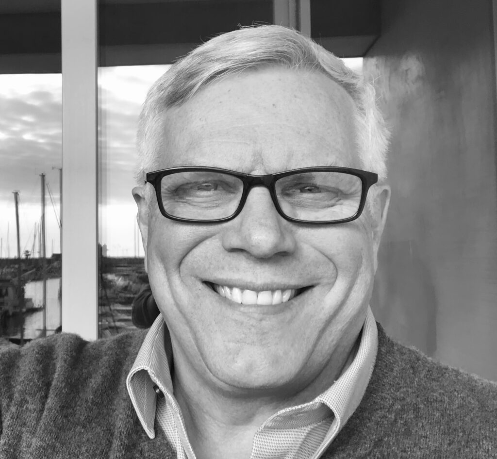 Black-and-white portrait of author John H. Thomas smiling warmly in front of a windowed backdrop with sailboats and reflections. Used to accompany his bio section for the Thunderbird Rising audiobook tour post on GinaRaeMitchell.com