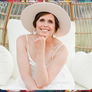 Author Paige Farrington, smiling warmly in a white dress and wide-brimmed hat, seated against a cozy woven backdrop