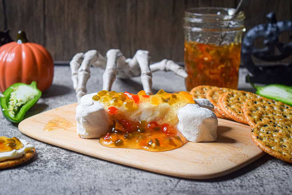 Cream cheese log shaped like a bone topped with spicy pepper jelly, surrounded by crackers and Halloween props including a skeleton hand, pumpkin, and jalapeño slices.