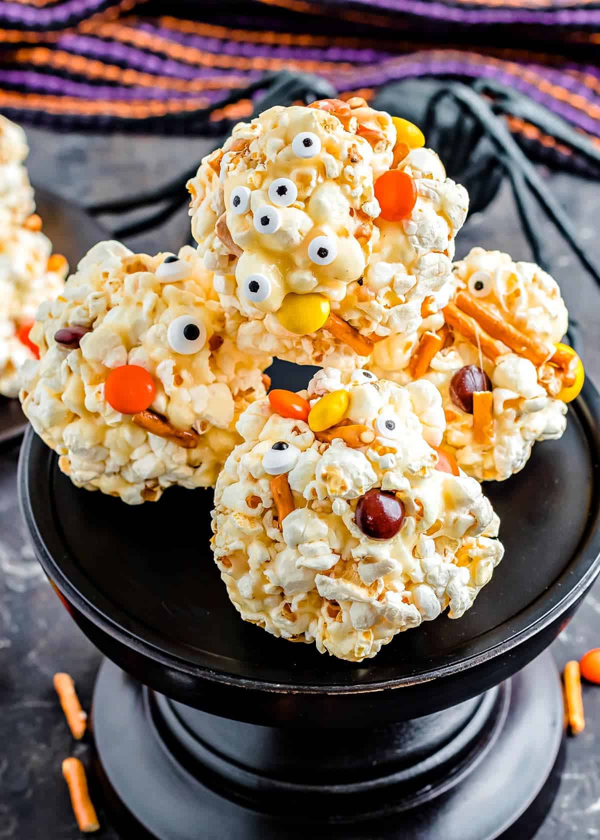 Halloween popcorn balls decorated with candy eyes, pretzel sticks, and colorful candies stacked on a black cake stand. Festive orange and purple striped ribbons in the background create a fun spooky party vibe.